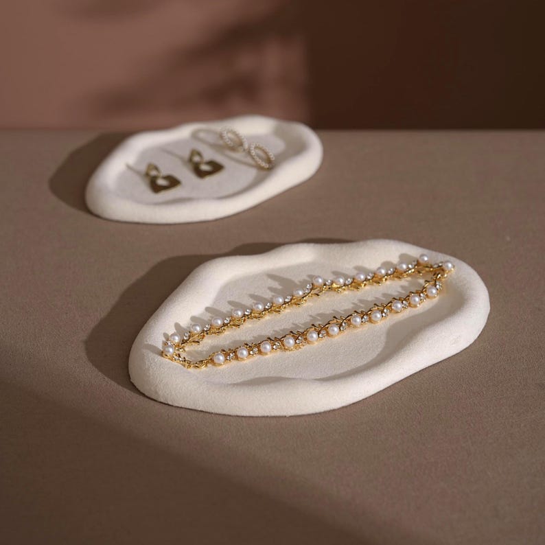 May include: Two off-white, organically shaped jewelry trays. One holds gold earrings, the other a gold necklace with pearls and small crystals. The trays sit on a neutral-toned surface, with a soft, diffused light.