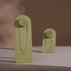 Op de afbeelding: Twee goudkleurige slangenkettingen gepresenteerd op boogvormige sieradenstandaards van saliegroen fluweel. De hogere standaard toont een langere, gelaagde ketting, terwijl de kleinere standaard een enkele, kortere ketting draagt met een gladde afwerking.