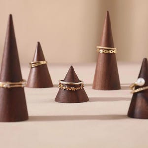 May include: Five dark brown wooden ring holders in a cone shape, each displaying a different gold ring. The rings vary in design, including bands with small stones and a ring with a single pearl. The holders are arranged on a light beige surface.