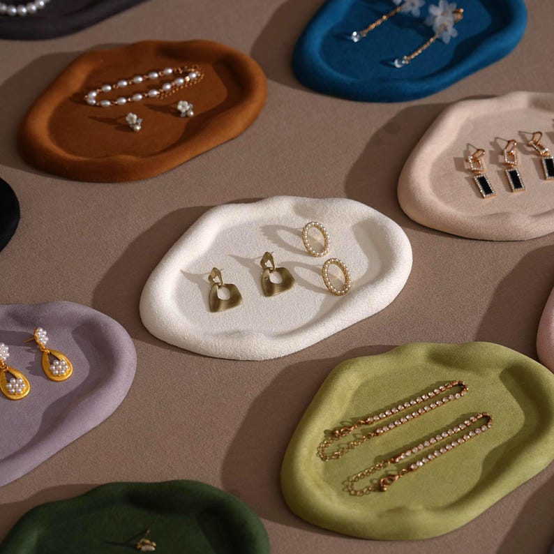 May include: An assortment of jewelry display trays in various colors, including brown, blue, green, and white. Each tray holds different pieces of jewelry, such as earrings, necklaces, and bracelets. The trays have an organic, irregular shape and a soft, textured surface.