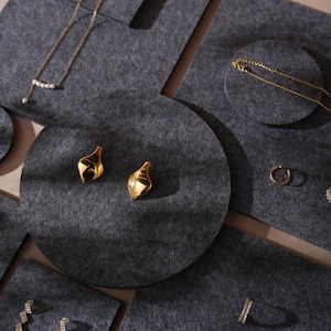 Op de afbeelding: Een set grijze vilten sieradenpresentatiekussens in verschillende vormen en maten. De kussens bevatten gouden oorbellen, een gouden ketting en een gouden ring.