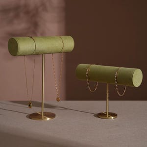 May include: Two jewelry display stands. Each stand has a cylindrical, olive green velvet bar atop a gold-colored metal stem and base. Gold necklaces hang from the bars. The stands are on a neutral surface against a brown background.