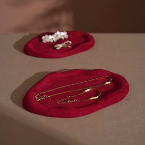 May include: Two red velvet jewelry trays. The top tray holds a white floral hair clip and a silver bow ring. The bottom tray displays two gold necklaces. The trays are irregularly shaped and sit on a neutral surface.