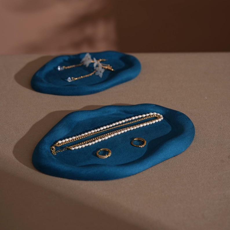 May include: Two teal-colored, organic-shaped jewelry trays. One tray holds gold earrings with white floral accents. The other tray displays a gold chain necklace with pearls, two gold rings, and a gold chain.