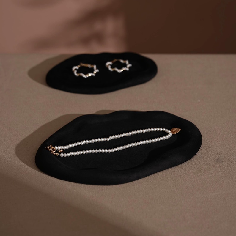 May include: Two black, organic-shaped trays. The top tray holds two gold-colored, cloud-shaped earrings. The bottom tray displays a white pearl necklace with a gold-colored clasp. The background is a neutral color.