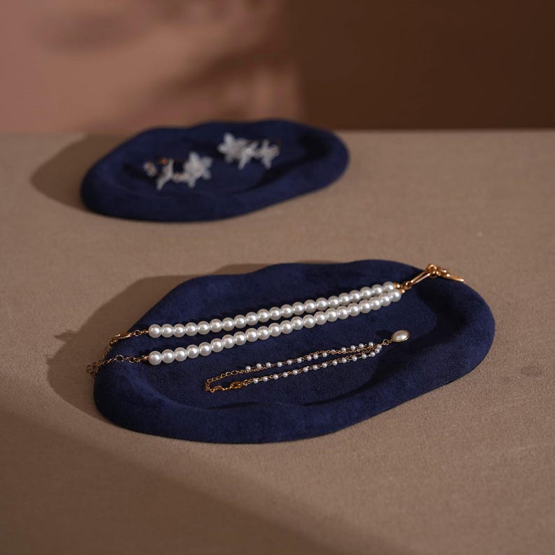 May include: Two navy blue felt jewelry trays. One tray holds a pearl necklace and a delicate pearl bracelet. The other tray holds a pair of floral earrings. The trays are on a neutral surface.