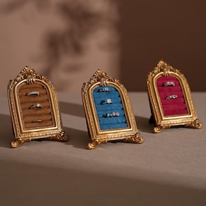 Könnte beinhalten: Drei verzierte, goldfarbene Ring-Ausstellungsständer. Jeder Ständer hat einen Samtinnenraum in Braun, Blau und Rot, der mehrere Ringe enthält. Die Ständer haben ein dekoratives, bogenförmiges Design und stehen auf einer neutralen Oberfläche.