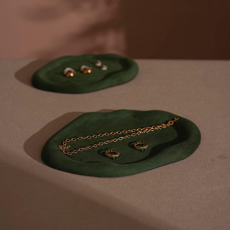 May include: Two dark green, organic-shaped jewelry trays. The bottom tray holds a gold necklace and two rings. The top tray holds three rings. The trays are on a neutral surface with a pink background.