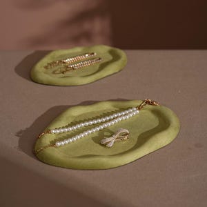 May include: Two olive-green velvet trays with jewelry. One tray holds two gold barrettes with clear stones. The other tray holds a pearl necklace and a gold bow-shaped hair clip with clear stones. The trays are on a neutral surface.