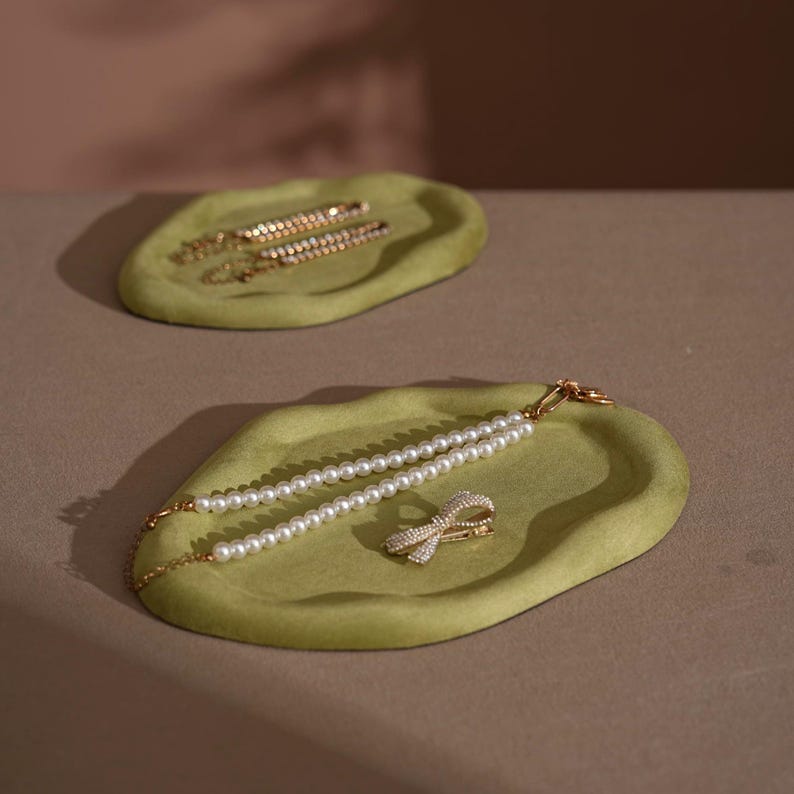 May include: Two olive-green velvet trays with jewelry. One tray holds two gold barrettes with clear stones. The other tray holds a pearl necklace and a gold bow-shaped hair clip with clear stones. The trays are on a neutral surface.