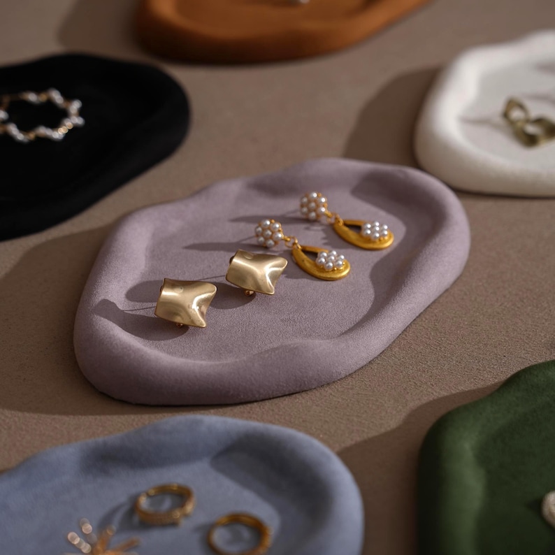 May include: A collection of small, irregularly shaped trays in various colors, including black, brown, and green. The trays hold jewelry, such as gold earrings with pearl accents and gold rings. The trays are displayed on a neutral surface.