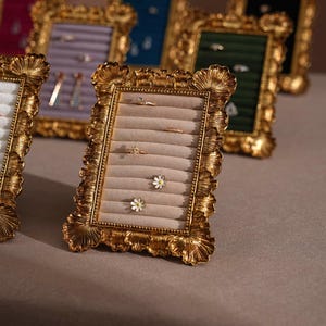 Könnte beinhalten: Goldfarbene Schmuckdisplays mit Rahmen und beigem Samtinterieur. Die Rahmen haben verzierte, strukturierte Kanten. Mehrere Ringe und ohrringförmige Ohrringe werden ausgestellt. Weitere Rahmen mit unterschiedlich farbigen Samtinterieurs sind im Hintergrund zu sehen.