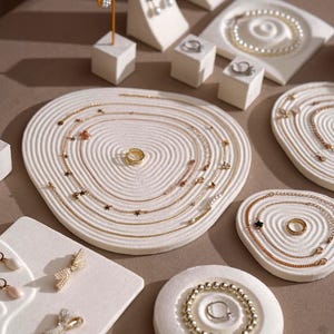 Könnte beinhalten: Eine Auswahl an goldenen Halsketten, Ringen, Ohrringen und Armbändern, präsentiert auf weißen, strukturierten Schmuckhaltern im Zen-Garten-Stil. Der Schmuck umfasst zarte Ketten mit Stern- und Perlenakzenten.