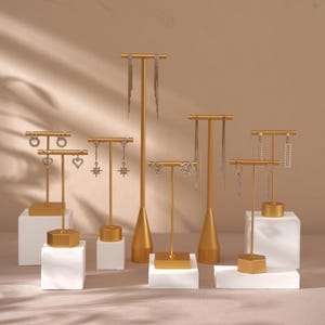 May include: Gold earring display stands with various styles of earrings. The stands have a gold metal base and a gold metal rod with a T-shaped top. The earrings are made of silver metal and have different designs, including stars, hearts, and circles.