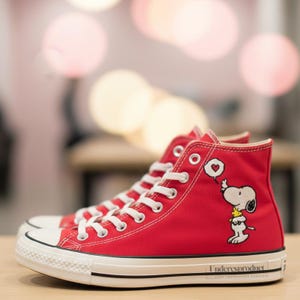 Puede incluir: Zapatillas altas rojas con cordones y suelas blancas. Las zapatillas presentan un diseño de dibujos animados de Snoopy y Woodstock en el lateral. Las suelas tienen una raya negra y el texto "Underesproduct" está impreso en el lateral.