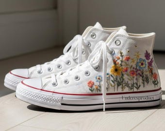 Könnte beinhalten: Weiße High-Top-Sneaker mit farbenfroher Blumenstickerei. Die Sneaker haben weiße Schnürsenkel, einen roten Streifen auf der Sohle und den Text "Underesproduct" an der Seite.