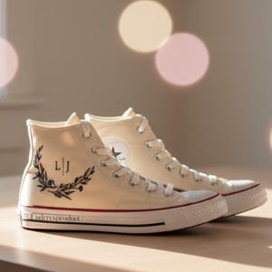 May include: Cream-colored high-top sneakers with white laces and a red stripe on the sole. The sneakers feature a black laurel wreath design with the initials "L|J" and the text "Underesproduct" on the side.