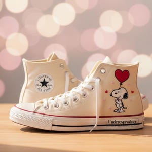 May include: Cream-coloured high-top trainers with a Snoopy graphic holding a heart-shaped balloon. The trainers have white laces, a black star logo, and a red stripe on the sole. The text "Underesproduct" is printed on the side.