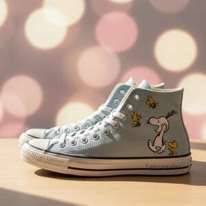Puede incluir: Zapatillas altas de color azul claro con un diseño de Snoopy y Woodstock. Las zapatillas tienen cordones blancos, una puntera de goma blanca y una raya negra a lo largo de la suela. Los zapatos están sobre una superficie de madera.