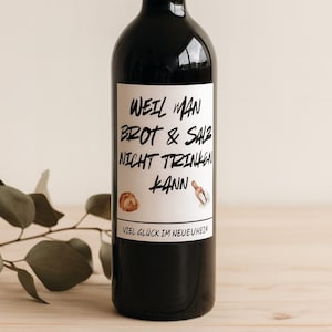 Könnte beinhalten: Eine schwarze Weinflasche mit einem weißen Etikett. Das Etikett trägt schwarzen Text, darunter "WEIL MAN BROT & SALZ NICHT TRINKEN KANN" und "VIEL GLÜCK IM NEUEUHEIN".
