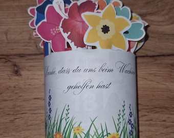 May include: A cylindrical paper gift container with a floral design and the German text "Danke, dass du uns beim Wachsen geholfen hast." The container is decorated with colorful paper flowers and green grass.