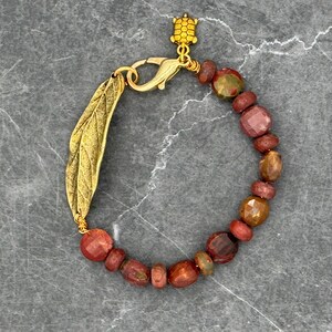 Puede incluir: Una pulsera de cuentas con cuentas rojas, marrones y doradas. Tiene un dije de hoja dorada y un dije de tortuga. El cierre también es dorado. La pulsera está sobre una superficie gris.