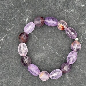 Könnte beinhalten: Ein Armband mit ovalen Amethyststeinen in Lila-, Lavendel- und transparenten Farbtönen. Die Perlen sind mit einem silberfarbenen Metallverschluss aufgefädelt. Das Armband wird auf einer grauen, strukturierten Oberfläche präsentiert.