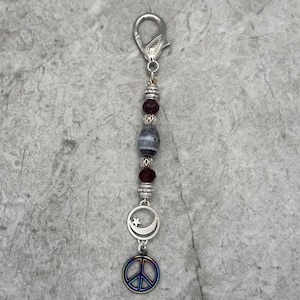 May include: A silver keychain with a lobster clasp, adorned with beads and charms. The design includes silver beads, dark red beads, a gray and white stone bead, and a crescent moon and star charm. A rainbow peace sign charm hangs at the bottom.