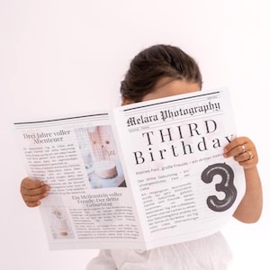 Puede incluir: Una persona sostiene un periódico con el titular "THIRD BIRTHDAY" en letras grandes y en negrita. El número "3" se muestra de forma destacada. El periódico también presenta texto en alemán y una foto de un pastel.