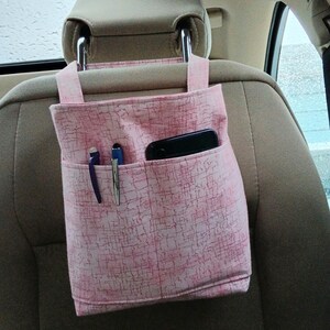 K&ouml;nnte beinhalten: Rosa Stoff-Auto-Organizer mit mehreren Taschen, der an einer Kopfst&uuml;tze befestigt ist. Der Organizer enth&auml;lt ein schwarzes Smartphone und zwei Stifte. Der Stoff hat ein dezentes, strukturiertes Muster.
