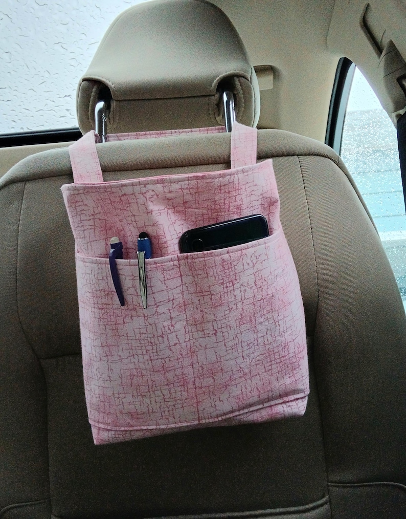 K&ouml;nnte beinhalten: Rosa Stoff-Auto-Organizer mit mehreren Taschen, der an einer Kopfst&uuml;tze befestigt ist. Der Organizer enth&auml;lt ein schwarzes Smartphone und zwei Stifte. Der Stoff hat ein dezentes, strukturiertes Muster.