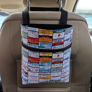 K&ouml;nnte beinhalten: Ein Autositz-Organizer mit einem Nummernschildmuster. Der Stoff zeigt eine Collage farbenfroher Nummernschilder aus verschiedenen Bundesstaaten, darunter New York, Montana und Louisiana. Er hat schwarze Aufh&auml;ngeschlaufen und eine schwarze Einfassung.