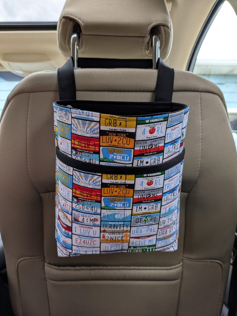 K&ouml;nnte beinhalten: Ein Autositz-Organizer mit einem Nummernschildmuster. Der Stoff zeigt eine Collage farbenfroher Nummernschilder aus verschiedenen Bundesstaaten, darunter New York, Montana und Louisiana. Er hat schwarze Aufh&auml;ngeschlaufen und eine schwarze Einfassung.