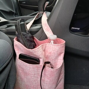 K&ouml;nnte beinhalten: Ein rosa Stoff-Auto-Organizer mit mehreren Taschen, der am Schalthebel h&auml;ngt. Der Organizer enth&auml;lt einen schwarzen Regenschirm, eine Wasserflasche, ein Smartphone und eine Sonnenbrille. Der Stoff hat ein subtiles, strukturiertes Muster.