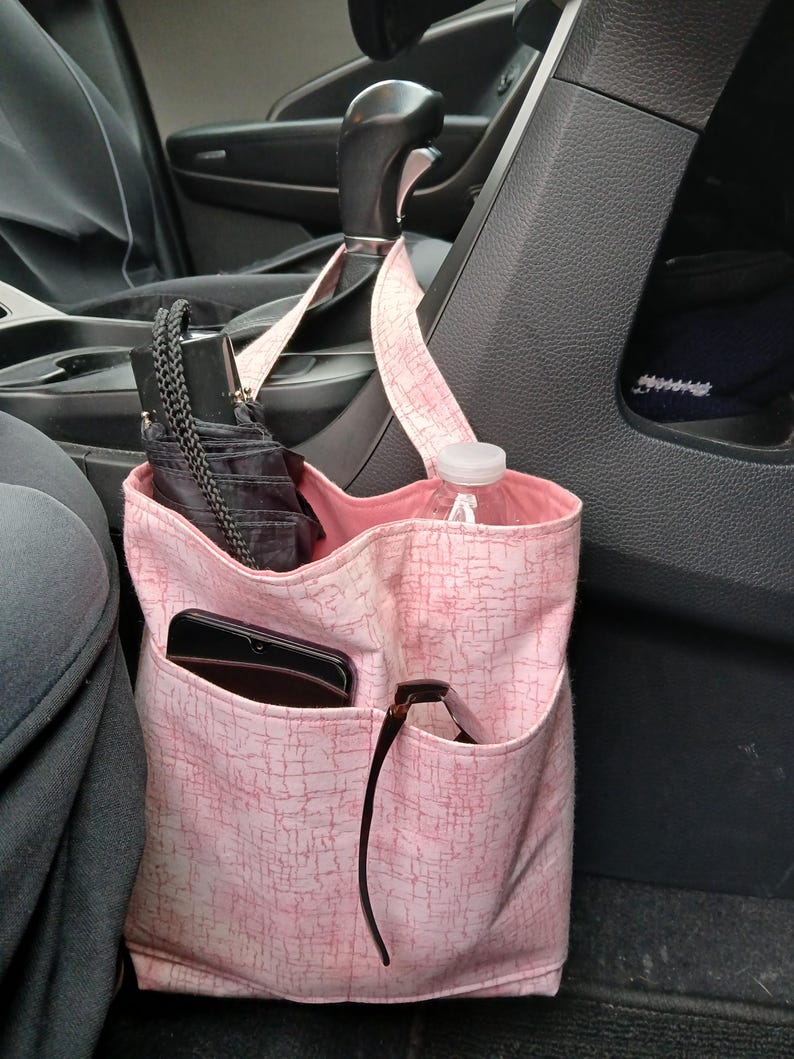 K&ouml;nnte beinhalten: Ein rosa Stoff-Auto-Organizer mit mehreren Taschen, der am Schalthebel h&auml;ngt. Der Organizer enth&auml;lt einen schwarzen Regenschirm, eine Wasserflasche, ein Smartphone und eine Sonnenbrille. Der Stoff hat ein subtiles, strukturiertes Muster.
