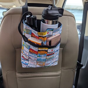 K&ouml;nnte beinhalten: Ein Autositz-Organizer mit einem Nummernschildmuster. Der Organizer h&auml;ngt an der Kopfst&uuml;tze und hat mehrere Taschen mit einer schwarzen Wasserflasche, einer wei&szlig;-schwarzen Wasserflasche, einem Telefon und anderen Gegenst&auml;nden. Der Hintergrund ist ein Autoinnenraum.