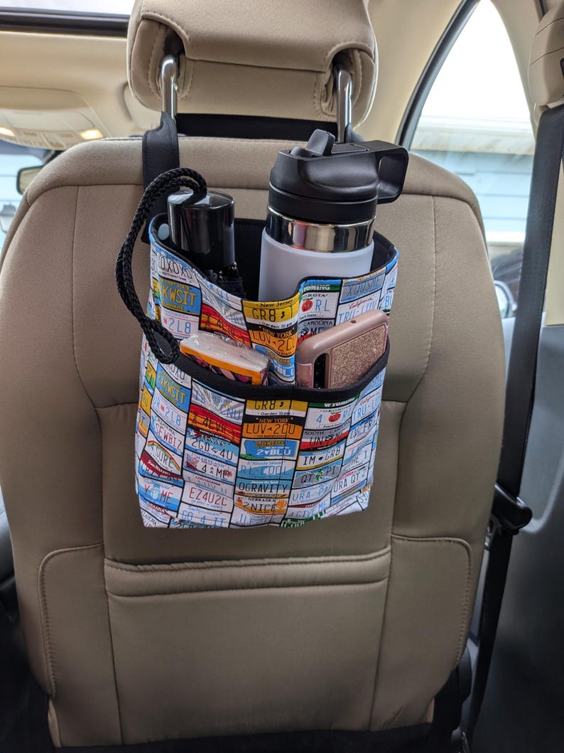 K&ouml;nnte beinhalten: Ein Autositz-Organizer mit einem Nummernschildmuster. Der Organizer h&auml;ngt an der Kopfst&uuml;tze und hat mehrere Taschen mit einer schwarzen Wasserflasche, einer wei&szlig;-schwarzen Wasserflasche, einem Telefon und anderen Gegenst&auml;nden. Der Hintergrund ist ein Autoinnenraum.