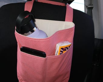 May include: A pink car seat organizer with white polka dots, hanging from a car headrest. It holds a water bottle, a smartphone, and a packet of tissues. The organizer has two pockets and a main compartment.