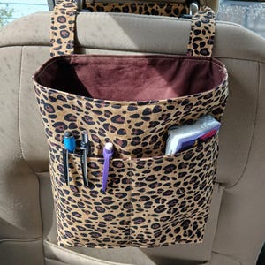 K&ouml;nnte beinhalten: Ein Autositz-Organizer mit braunem und schwarzem Leopardenmuster. Der Organizer hat ein gro&szlig;es offenes Fach mit braunem Innenraum und eine Vordertasche mit Stiften und Taschent&uuml;chern. Gurte werden an der Kopfst&uuml;tze befestigt.