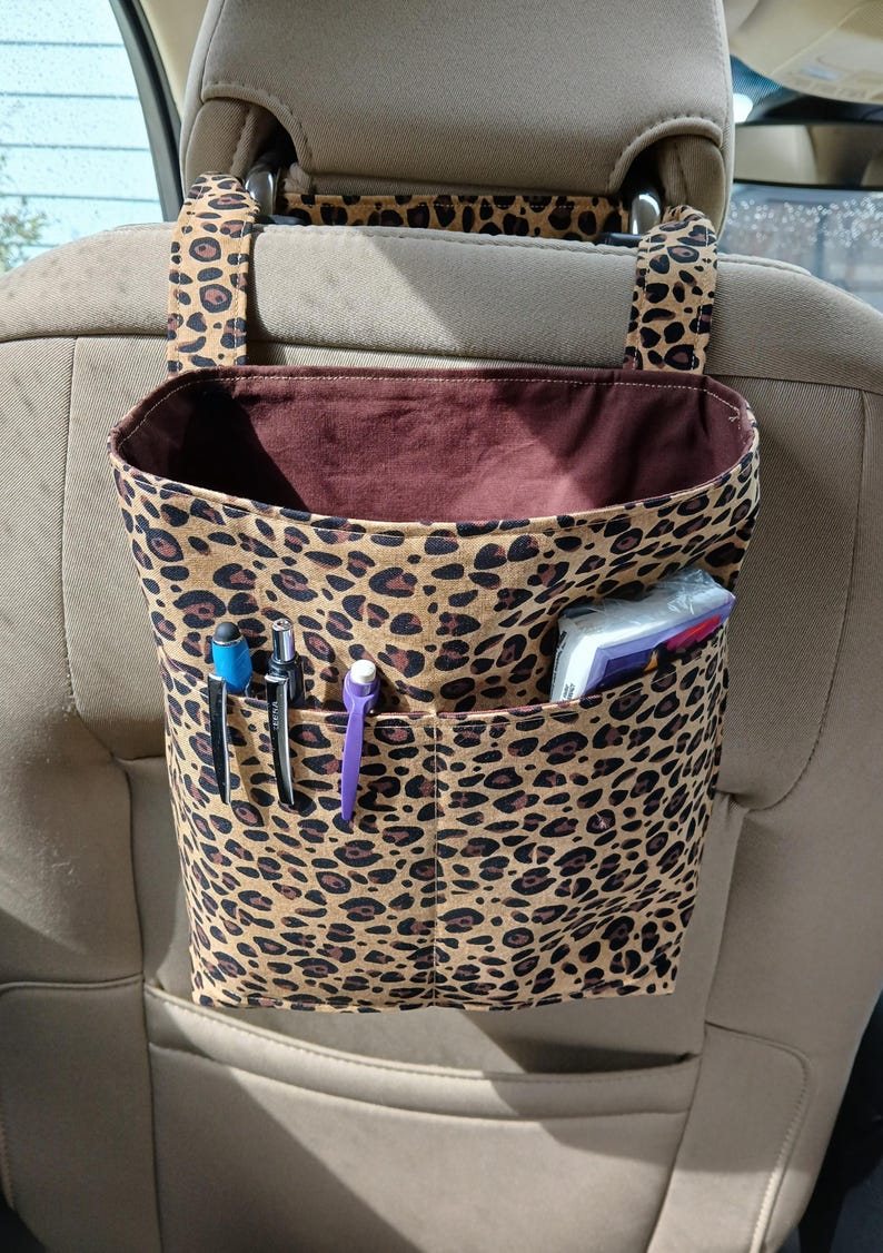 K&ouml;nnte beinhalten: Ein Autositz-Organizer mit braunem und schwarzem Leopardenmuster. Der Organizer hat ein gro&szlig;es offenes Fach mit braunem Innenraum und eine Vordertasche mit Stiften und Taschent&uuml;chern. Gurte werden an der Kopfst&uuml;tze befestigt.