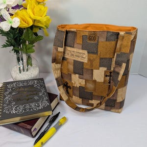 May include: A fabric tote bag featuring a patchwork pattern in shades of brown and tan, decorated with cross symbols, deer silhouettes, and the text Strong and courageous for the Lord your God. The bag has an orange interior lining and two straps.
