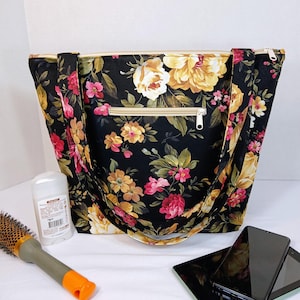 May include: Black tote bag with a floral pattern featuring yellow, pink, and red flowers. The bag has a zipper closure and long straps. A deodorant stick, a hairbrush, a smartphone, and a tablet are next to the bag.