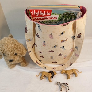 May include: A cream-colored tote bag with a pink interior and handles, filled with Highlights Hidden Pictures books. The bag is decorated with small animal illustrations. Toy lions, a zebra, and a stuffed dog are in the foreground.