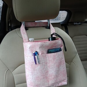 K&ouml;nnte beinhalten: Rosa Stoff-Auto-Organizer mit mehreren Taschen, der an einer Kopfst&uuml;tze befestigt ist. Der Organizer enth&auml;lt eine Wasserflasche, einen Stift, ein Telefon und eine schwarze Flasche. Der Stoff hat ein subtiles, strukturiertes Muster.