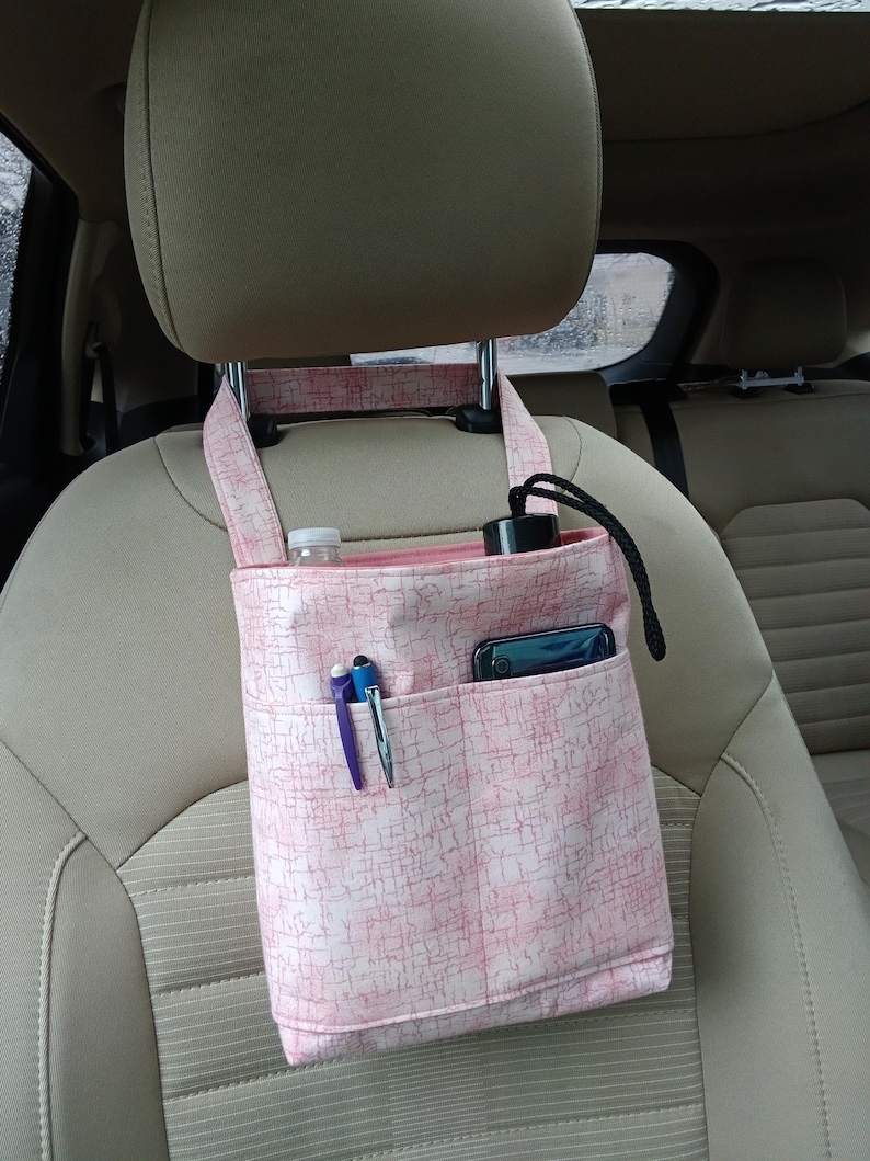 K&ouml;nnte beinhalten: Rosa Stoff-Auto-Organizer mit mehreren Taschen, der an einer Kopfst&uuml;tze befestigt ist. Der Organizer enth&auml;lt eine Wasserflasche, einen Stift, ein Telefon und eine schwarze Flasche. Der Stoff hat ein subtiles, strukturiertes Muster.