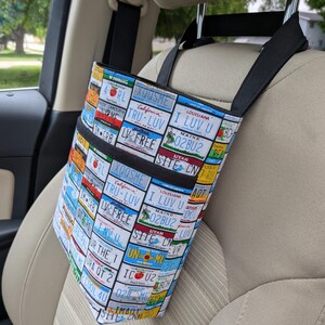 K&ouml;nnte beinhalten: Eine Auto-Organizer-Tasche mit einem farbenfrohen Nummernschildmuster. Die Tasche hat zwei schwarze Taschen und schwarze Tr&auml;ger, die &uuml;ber der Kopfst&uuml;tze h&auml;ngen. Die Tasche ist so konzipiert, dass sie Gegenst&auml;nde aufnehmen und diese w&auml;hrend der Fahrt organisiert halten.