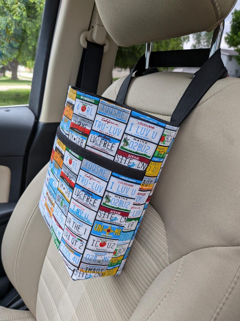 K&ouml;nnte beinhalten: Eine Auto-Organizer-Tasche mit einem farbenfrohen Nummernschildmuster. Die Tasche hat zwei schwarze Taschen und schwarze Tr&auml;ger, die &uuml;ber der Kopfst&uuml;tze h&auml;ngen. Die Tasche ist so konzipiert, dass sie Gegenst&auml;nde aufnehmen und diese w&auml;hrend der Fahrt organisiert halten.