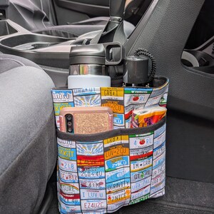 K&ouml;nnte beinhalten: Ein Stoff-Auto-Organizer mit mehreren Taschen, verziert mit bunten Nummernschildern. Der Organizer bietet Platz f&uuml;r eine Wasserflasche, ein Telefon und andere kleine Gegenst&auml;nde. Er ist f&uuml;r die Platzierung zwischen den Vordersitzen eines Fahrzeugs konzipiert.