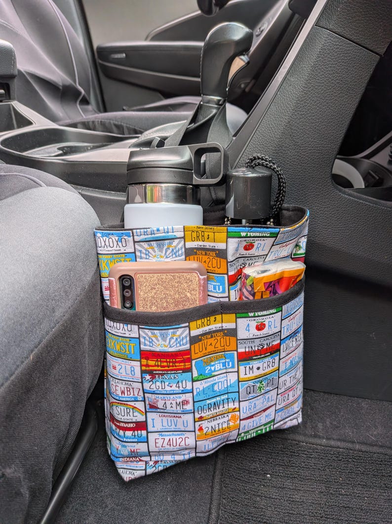 K&ouml;nnte beinhalten: Ein Stoff-Auto-Organizer mit mehreren Taschen, verziert mit bunten Nummernschildern. Der Organizer bietet Platz f&uuml;r eine Wasserflasche, ein Telefon und andere kleine Gegenst&auml;nde. Er ist f&uuml;r die Platzierung zwischen den Vordersitzen eines Fahrzeugs konzipiert.