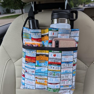 K&ouml;nnte beinhalten: Autositz-Organizer mit farbenfrohem Nummernschildmuster. Enth&auml;lt mehrere F&auml;cher mit einer schwarzen Wasserflasche, einem Edelstahlbecher, einem Telefon und anderen Gegenst&auml;nden. Der Organizer wird an der Kopfst&uuml;tze aufgeh&auml;ngt.